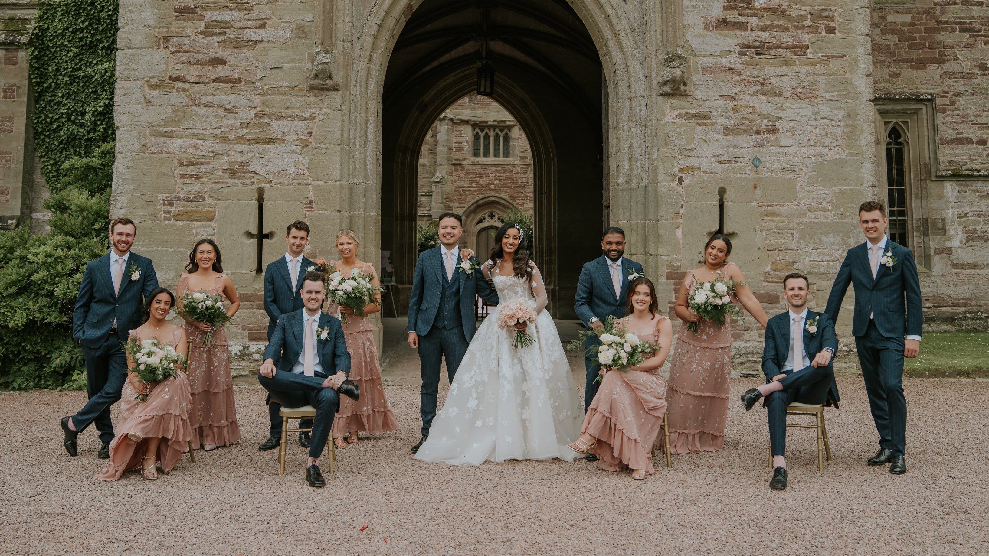 Wedding party group photo with bridesmaids holding white and green bouquets and groomsmen wearing white floral buttonholes at Hampton Court Castle's stone archway - Floral Design by Amaranté London.