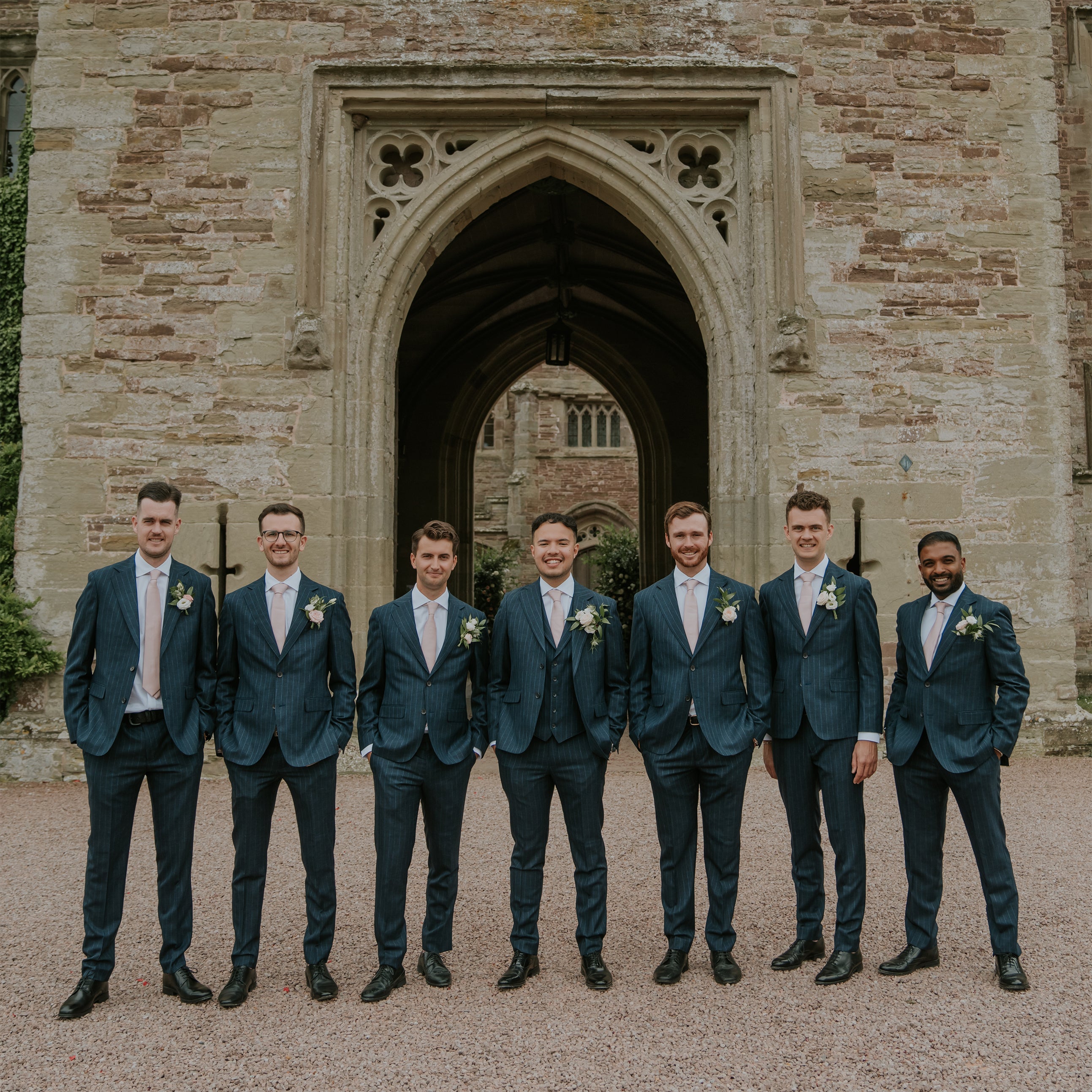 Groomsmen in navy suits with matching white and green floral buttonholes standing at Hampton Court Castle's ornate stone archway - Floral Design by Amaranté London.