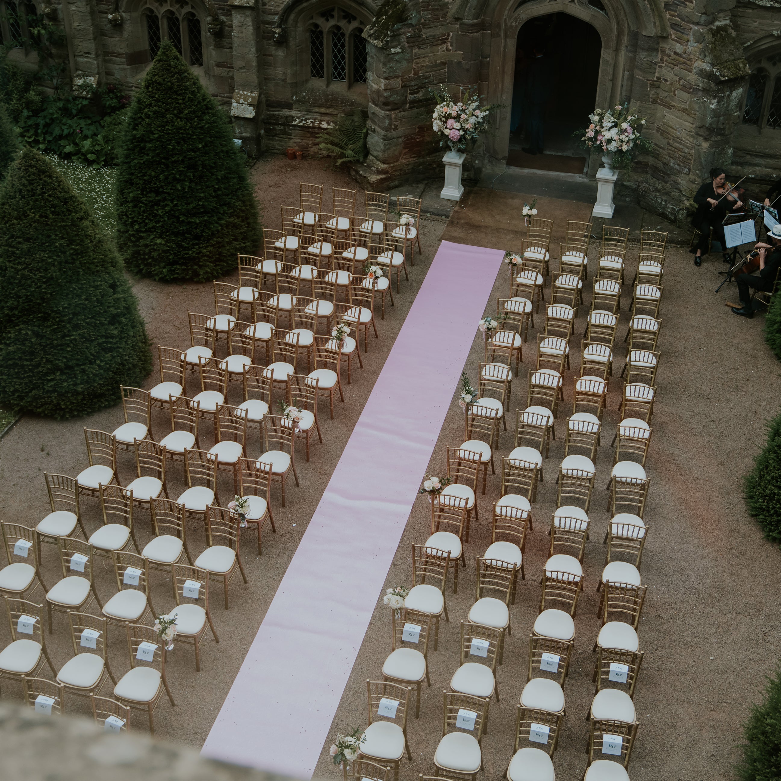 Aerial view of outdoor wedding ceremony setup with gold Chiavari chairs, pink aisle runner, and elegant floral arrangements on pedestals flanking Hampton Court Castle's historic stone courtyard - Floral Design by Amaranté London.