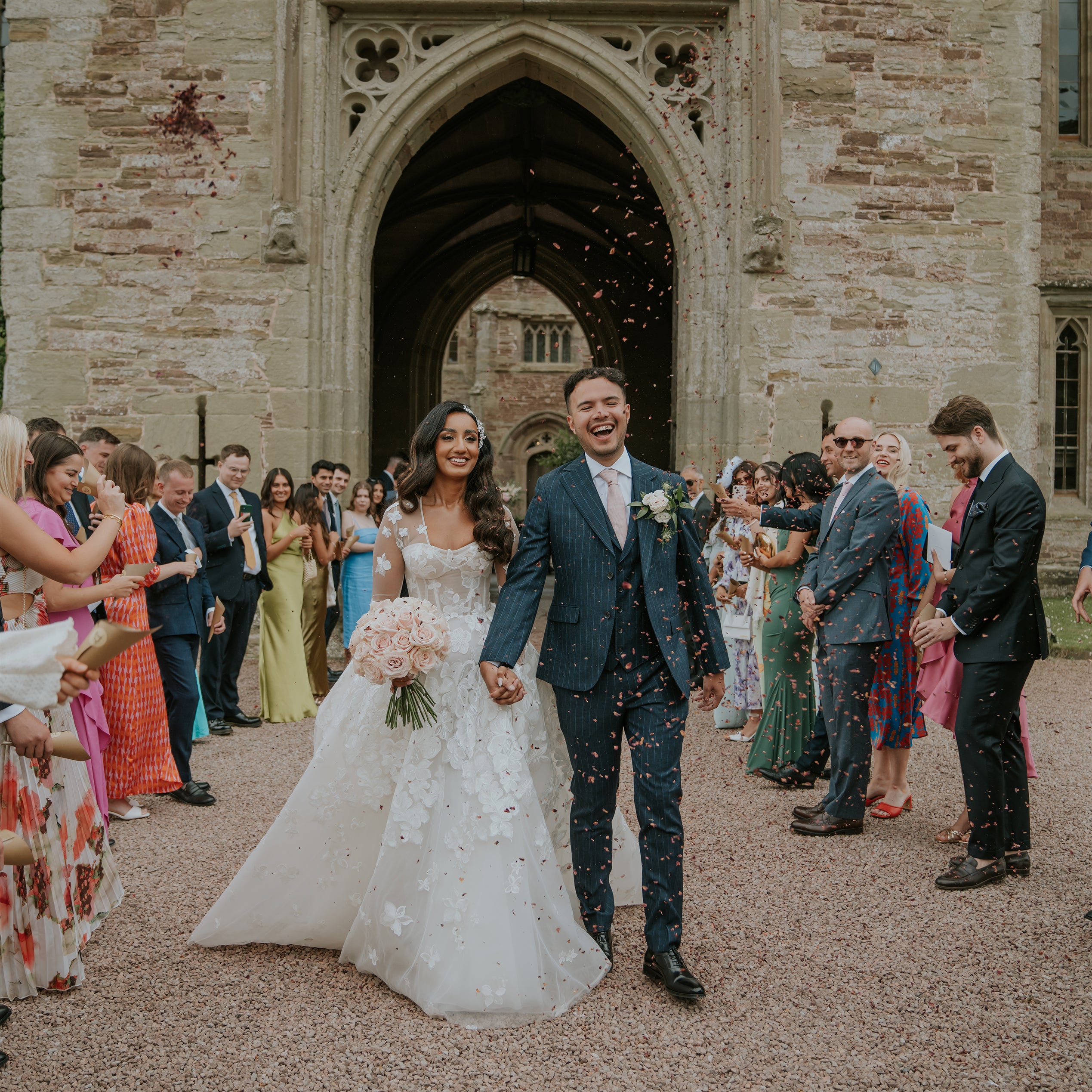 Newlyweds walking through a confetti shower, the bride holds a bridal bouquet of blush pink roses at Hampton Court Castle Wedding Venue