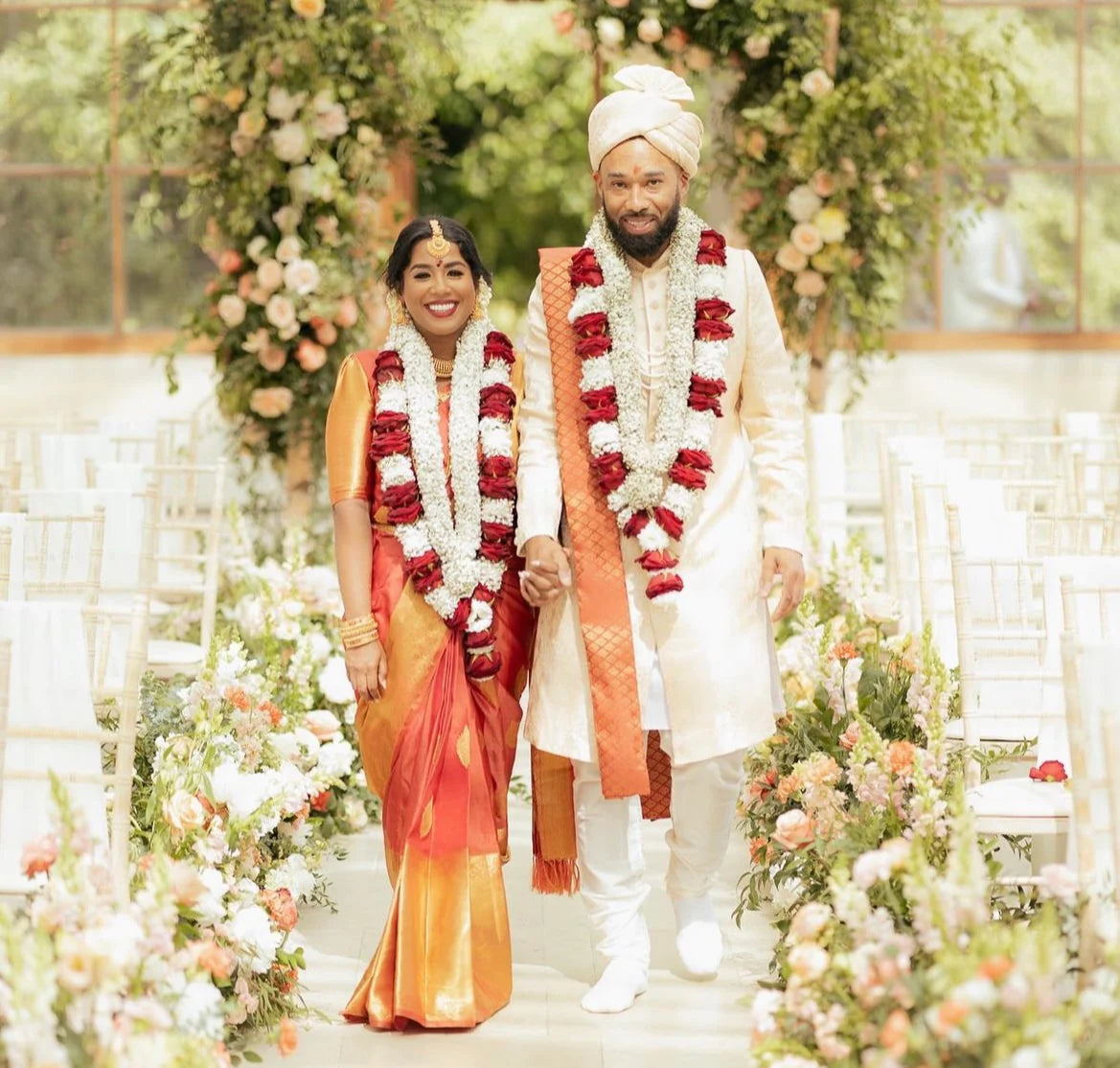 Hindu wedding ceremony at Kew Gardens with bride and groom walking through aisle lined with lush peach and white floral arrangements, beneath an elaborate floral arch with roses and trailing greenery in the conservatory - Amaranté London.