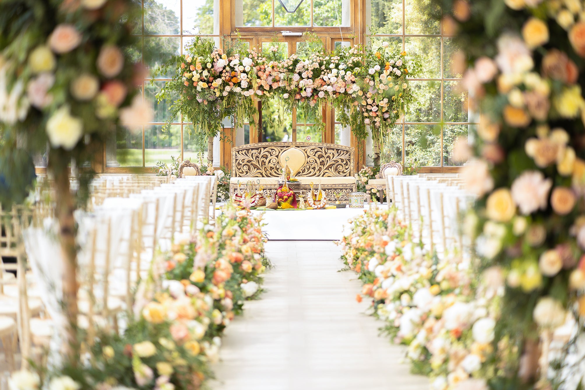 Spectacular floral ceremony arch at Kew Gardens with peach, pink, and white roses cascading with trailing greenery, complemented by matching floral arrangements lining the ceremony aisle in the light-filled conservatory - Amaranté London.