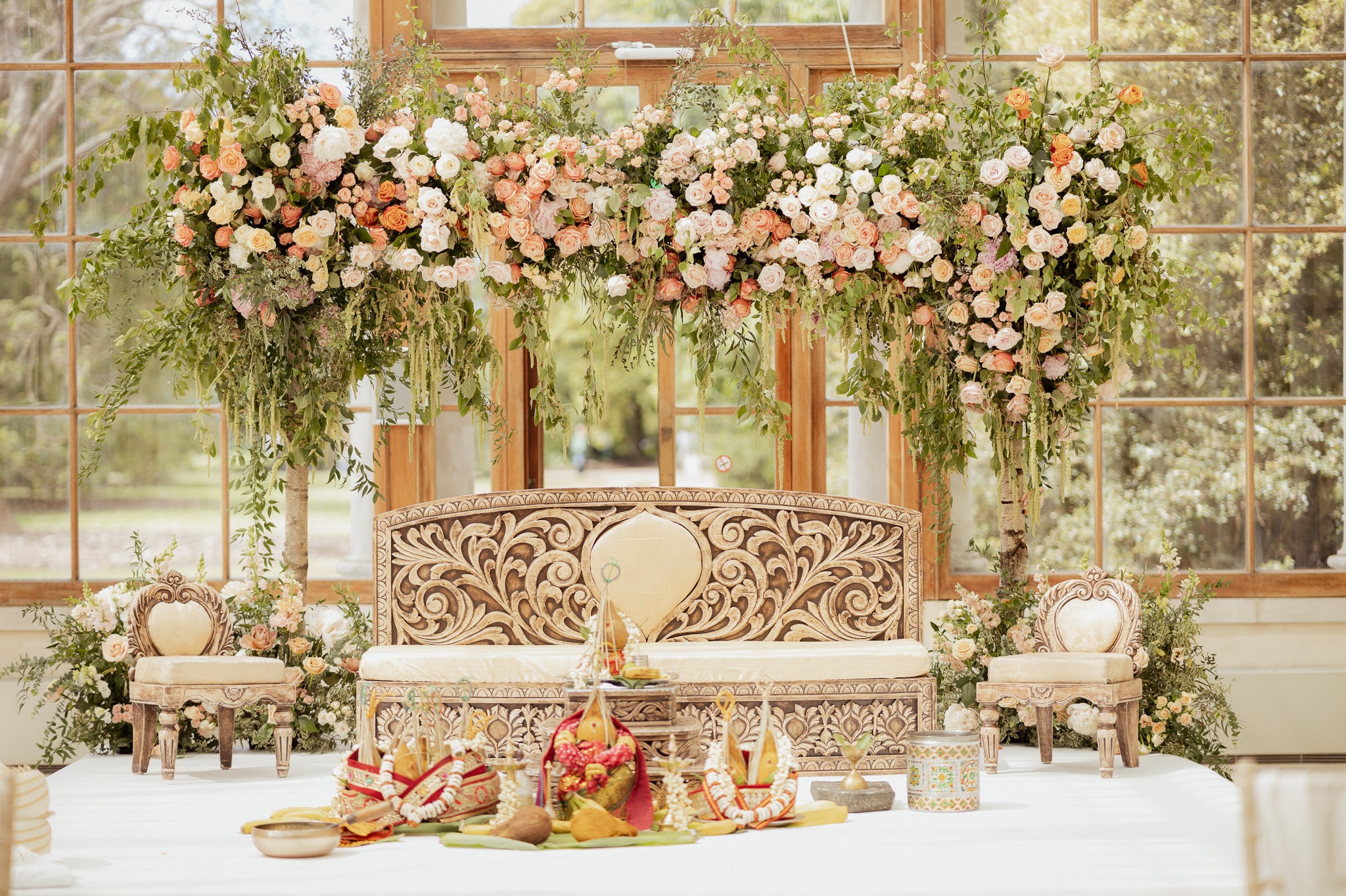 Elaborate Hindu wedding mandap at Kew Gardens featuring abundant floral arch with peach, coral, and white roses cascading with trailing eucalyptus and greenery, complemented by matching floral arrangements on an ornate carved backdrop and ceremonial seating in the sunlit conservatory - Amaranté London.