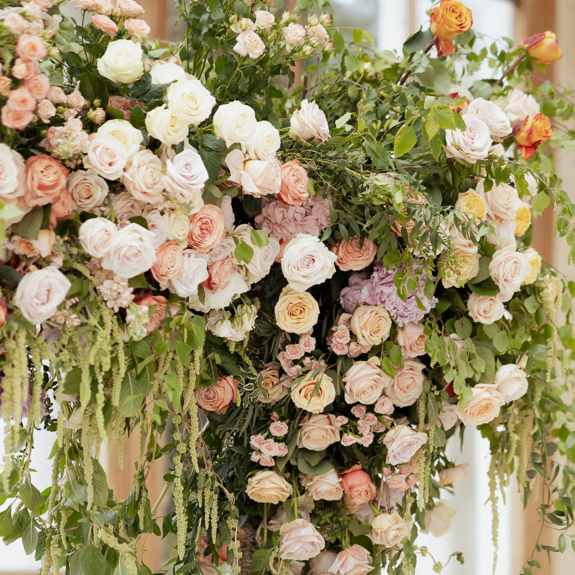 Luxurious floral arrangement at Kew Gardens with layered peach, blush, and white roses, mauve hydrangeas, coral accent blooms, and trailing amaranthus and greenery creating an abundant, textured display in the conservatory - Amaranté London.