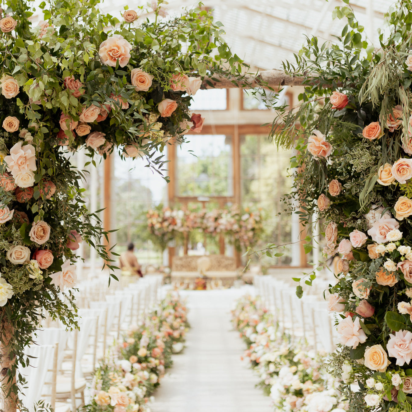 Organic floral ceremony arch at Kew Gardens, featuring peach and blush roses, mixed greenery including eucalyptus and olive branches, on a natural wooden beam, framing the view of matching floral aisle arrangements in a sunlit conservatory - Amaranté London.