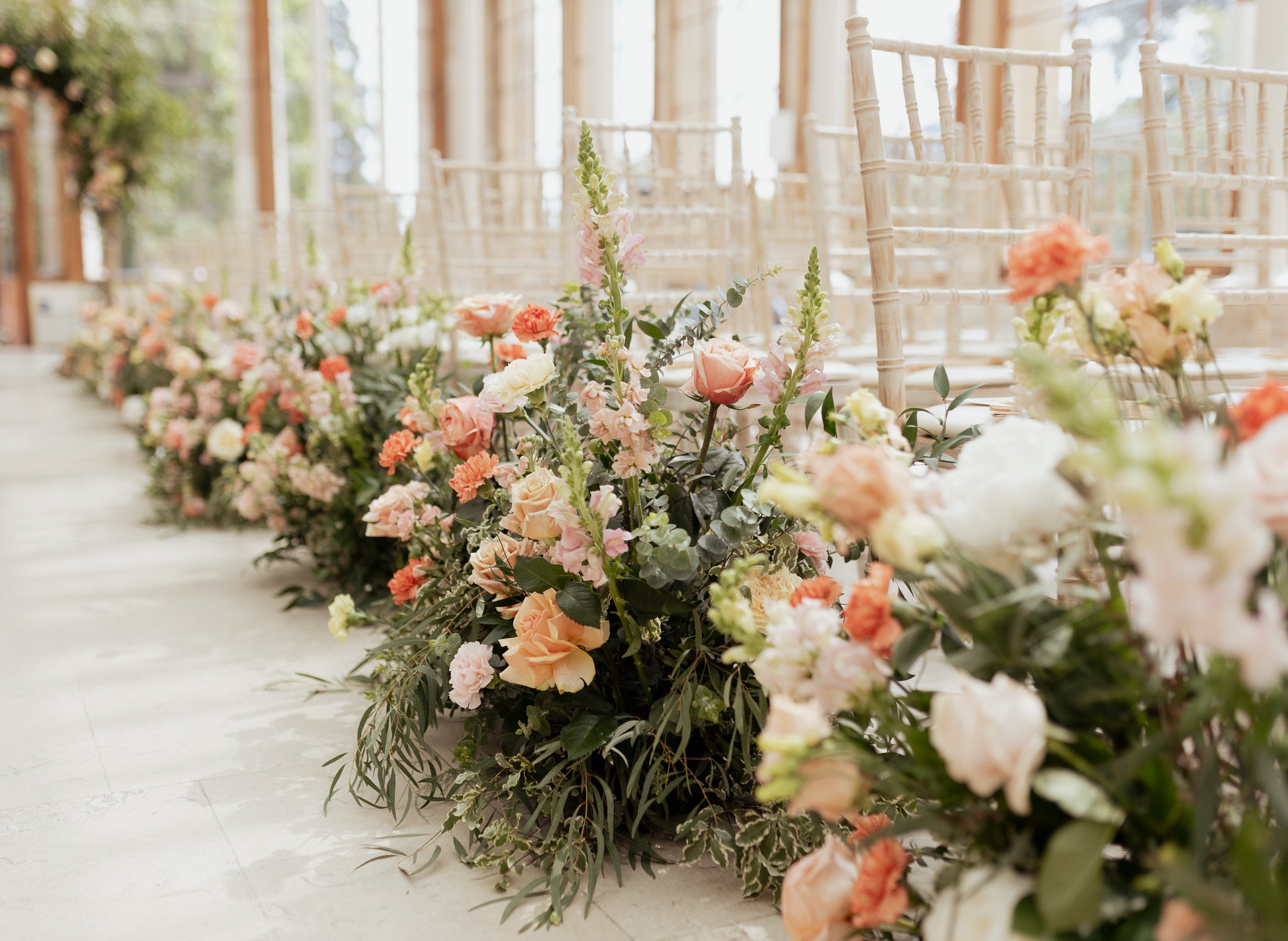 Romantic ceremony aisle at Kew Gardens, lined with organic floral arrangements featuring coral roses, peach blooms, white snapdragons, eucalyptus, and mixed greenery in natural clusters, along a white runner with limewashed Chiavari chairs in a light-filled conservatory - Amaranté London.