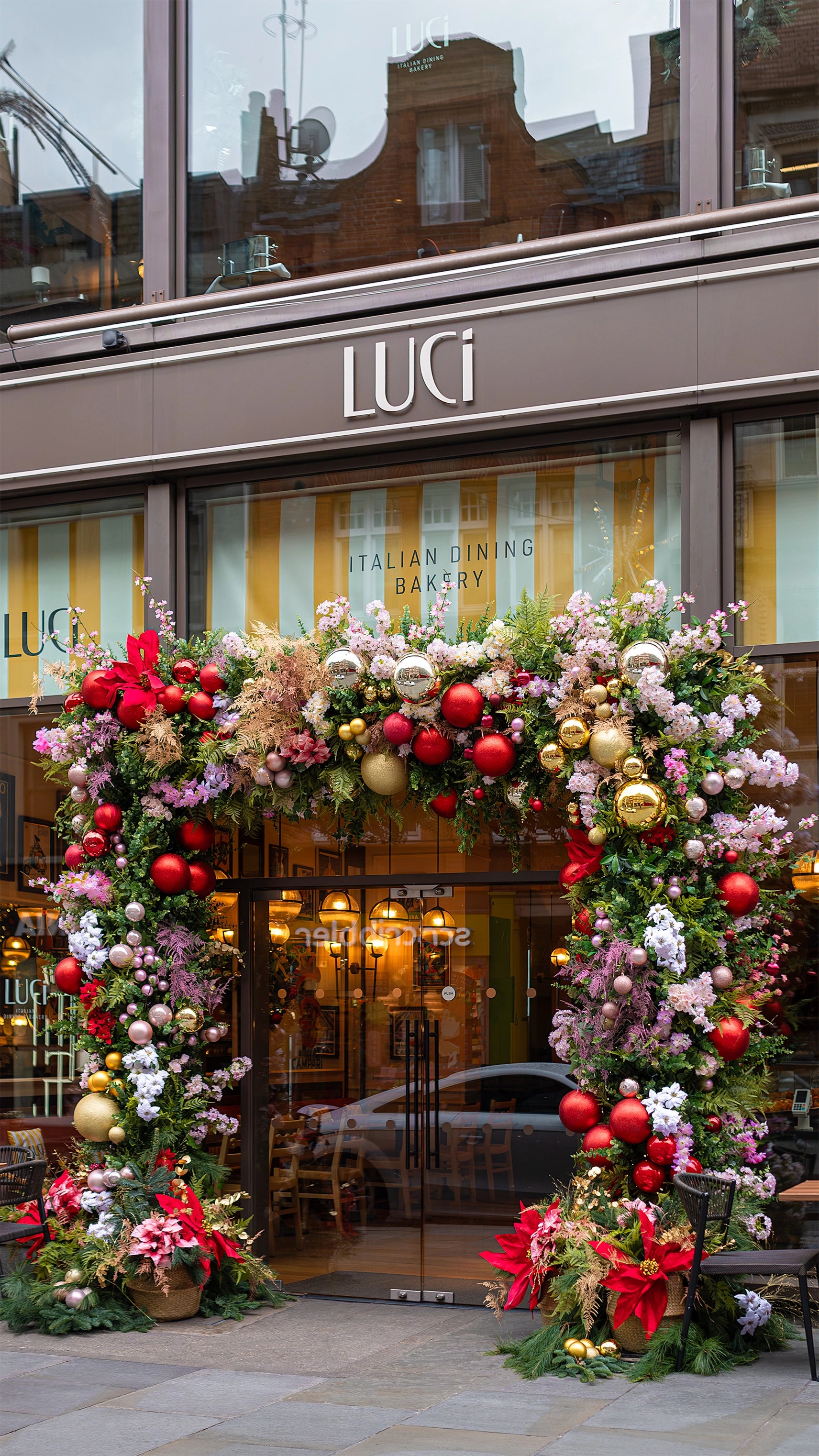 Festive Christmas floral archway installation with red and gold baubles, pink blooms, and evergreen foliage adorning restaurant entrance, showcasing Amaranté London's seasonal retail space transformations