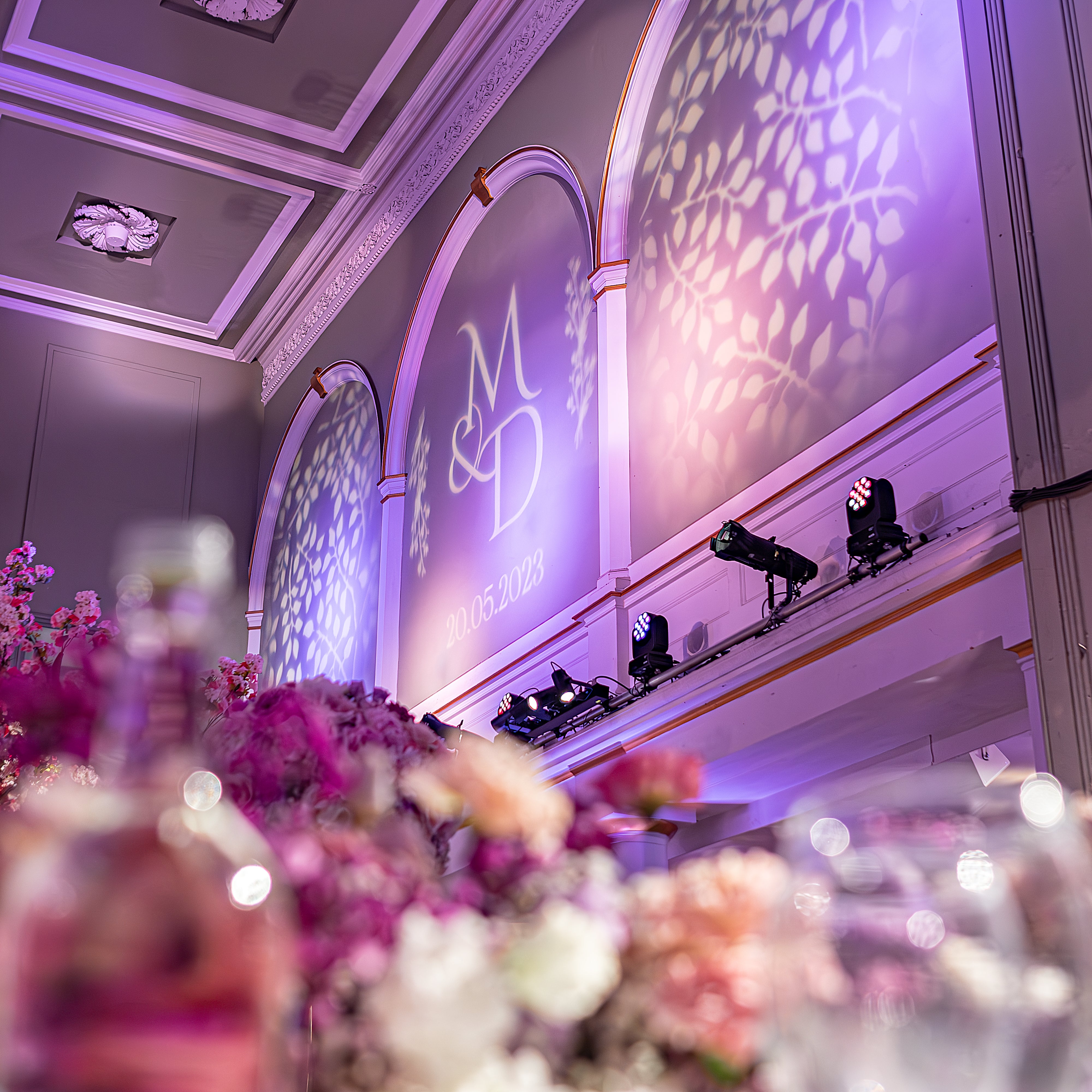 One Marylebone reception features vibrant pink and fuchsia floral arrangements in the foreground, custom monogram gobo lighting projection on neoclassical walls, and purple atmospheric uplighting that transforms the elegant Soane Hall architecture - Floral Design by Amaranté London.