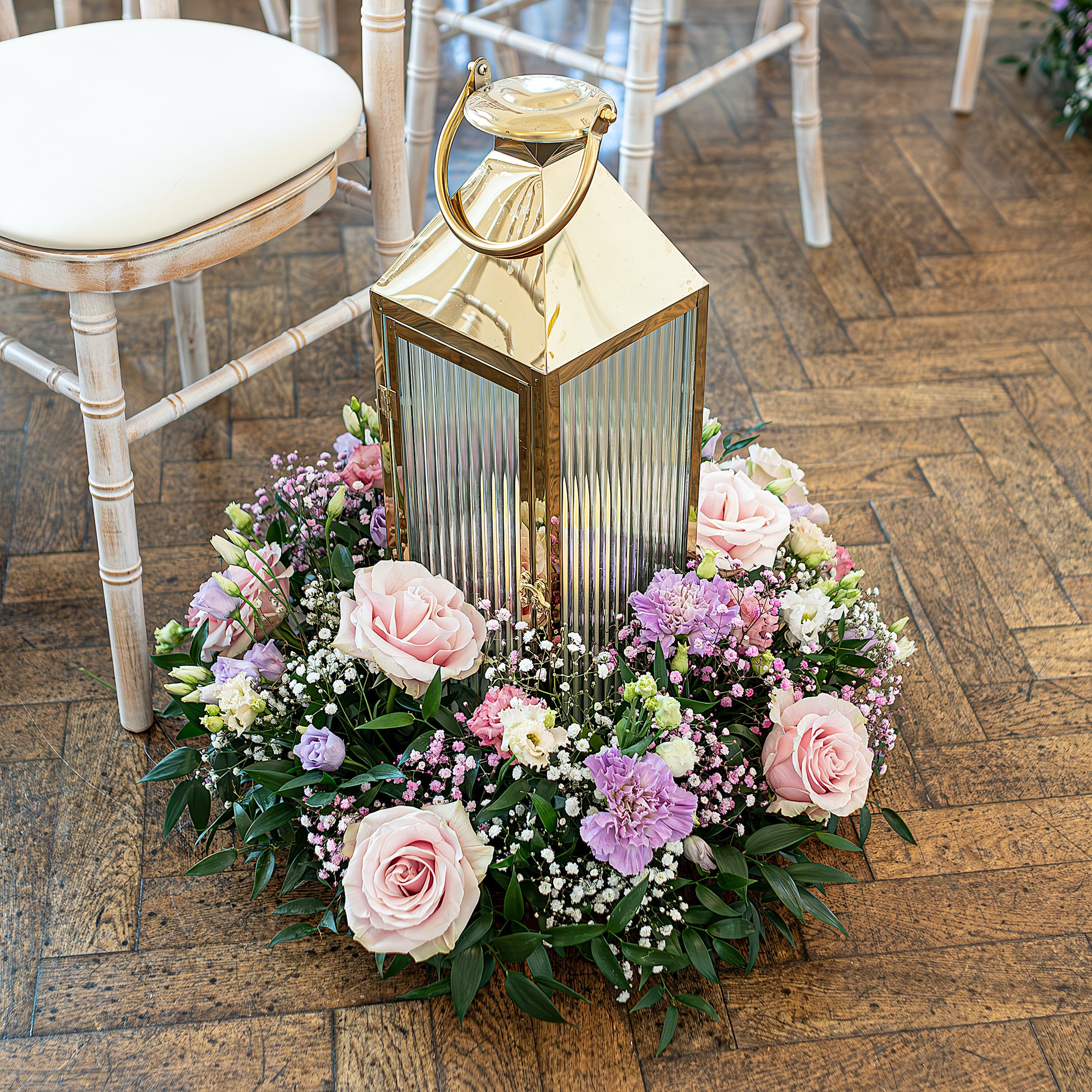 One Marylebone wedding venue aisle decoration features a gold lantern surrounded by a floral wreath of pink roses, purple scabiosa, white baby's breath, and lush greenery on a rustic wooden floor, beside a limewashed chiavari chair. Floral Design by Amaranté London.