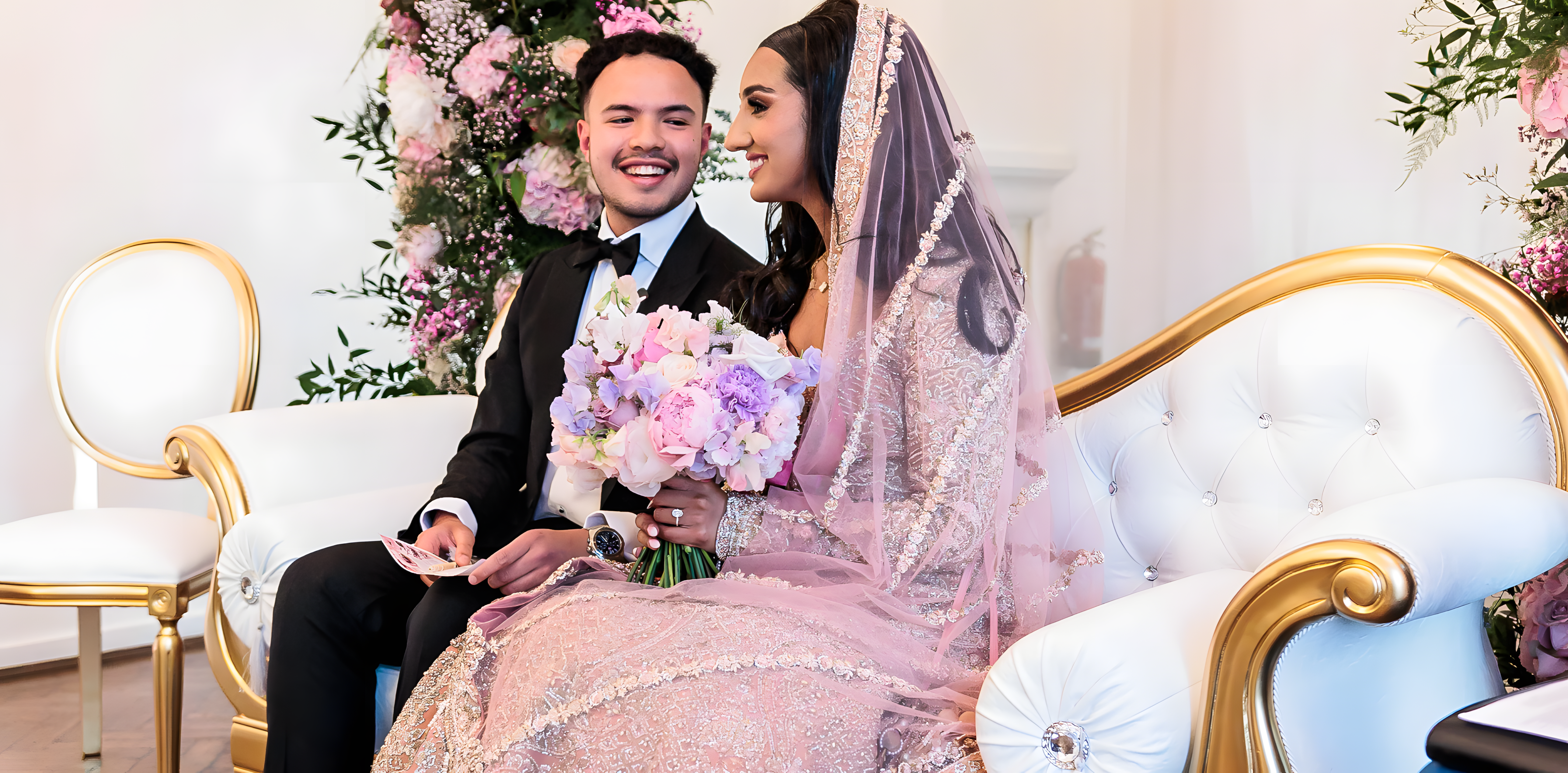 Bride holding luxury bridal bouquet of pink hydrangeas, lavender sweet peas, and white blooms at One Marylebone wedding venue, seated on ornate gold and white tufted sofa with matching floral arch backdrop featuring pink, purple, and white flowers with trailing greenery - Floral Design by Amaranté London.
