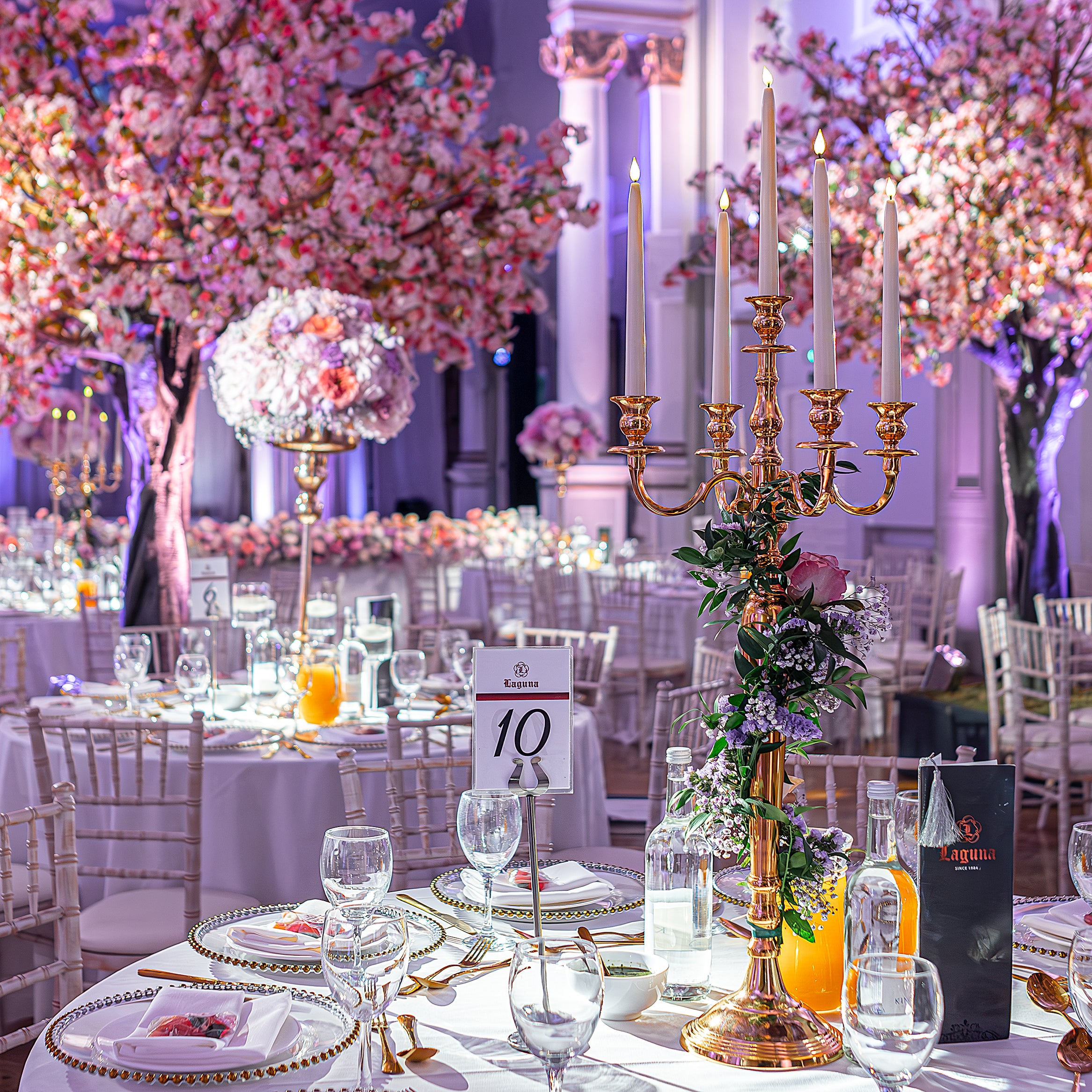 Whimsical One Marylebone wedding reception with tall gold candelabra centerpiece adorned with trailing purple wax flowers and greenery, cherry blossom trees with pink blooms, and elevated floral spheres of white hydrangeas and pink roses on gold stands creating a warm, cheerful atmosphere in an elegant gallery - Floral Design by Amaranté London.