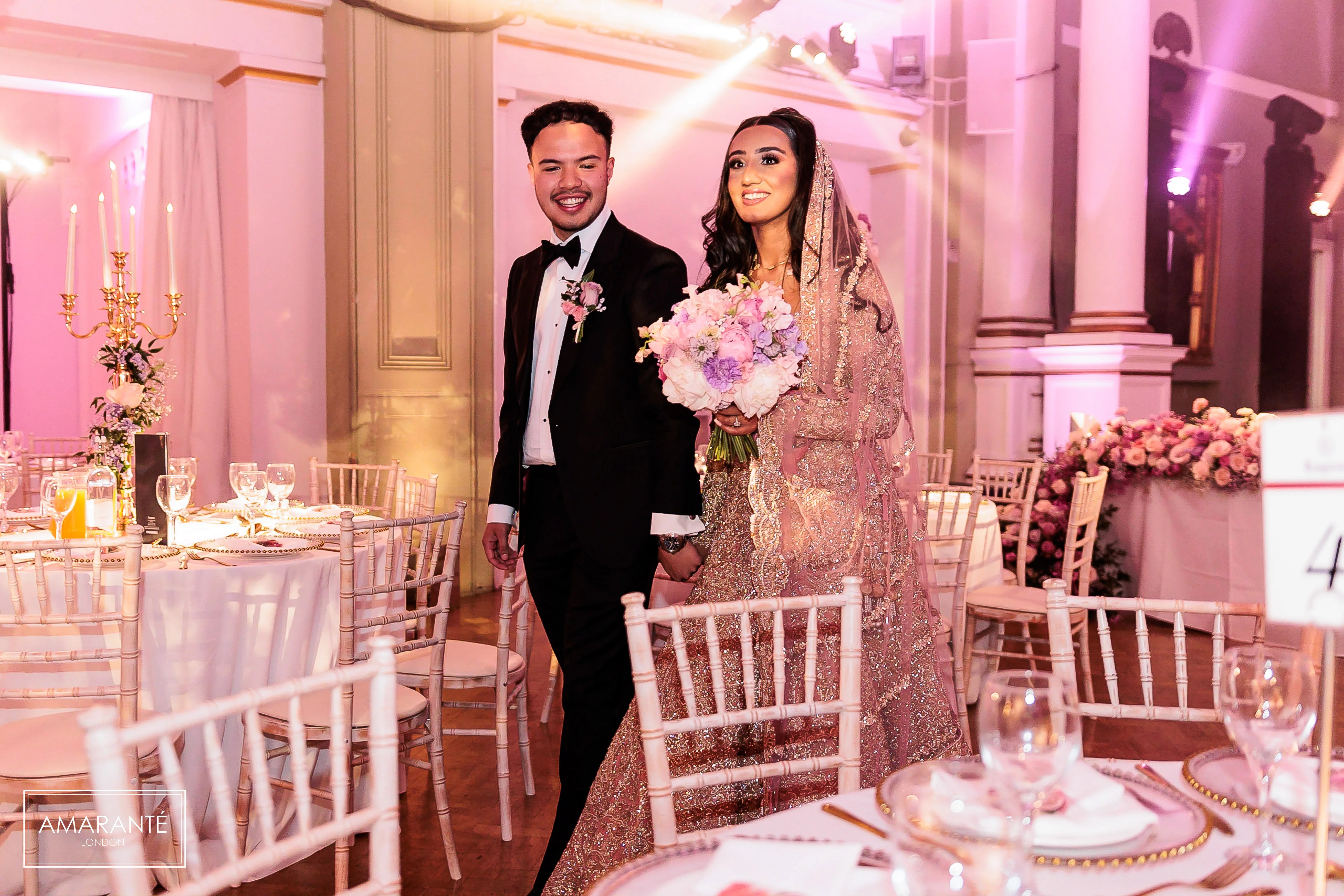Bride holding soft pink and lavender bridal bouquet at One Marylebone reception with tall gold candelabra adorned with trailing pink florals and lush pink floral arrangements visible in elegant neoclassical gallery space - Amaranté London.