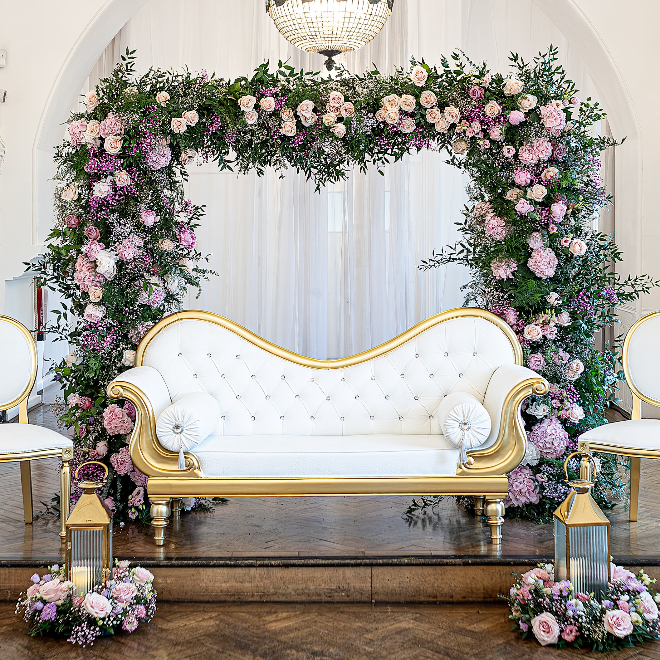 Luxurious floral arch at One Marylebone with pink hydrangeas, blush roses, purple wax flowers, and lush greenery framing an ornate gold and white tufted sofa, flanked by matching floral arrangements on gold pedestals in an elegant neoclassical gallery - Floral Design by Amaranté London.