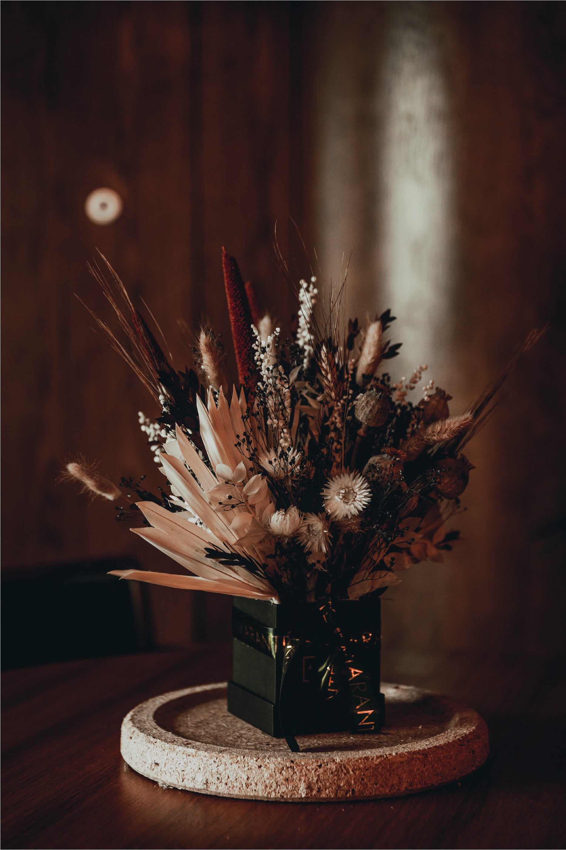 This image captures the intricate detail of this bespoke floral arrangement designed and produced by Amaranté London, a lush composition with striking red spikes, soft white flowers, and textural foliage, all contained in a dark, elegant box vase resting on a cork display in the warm, wood-toned ambience of a London event venue.