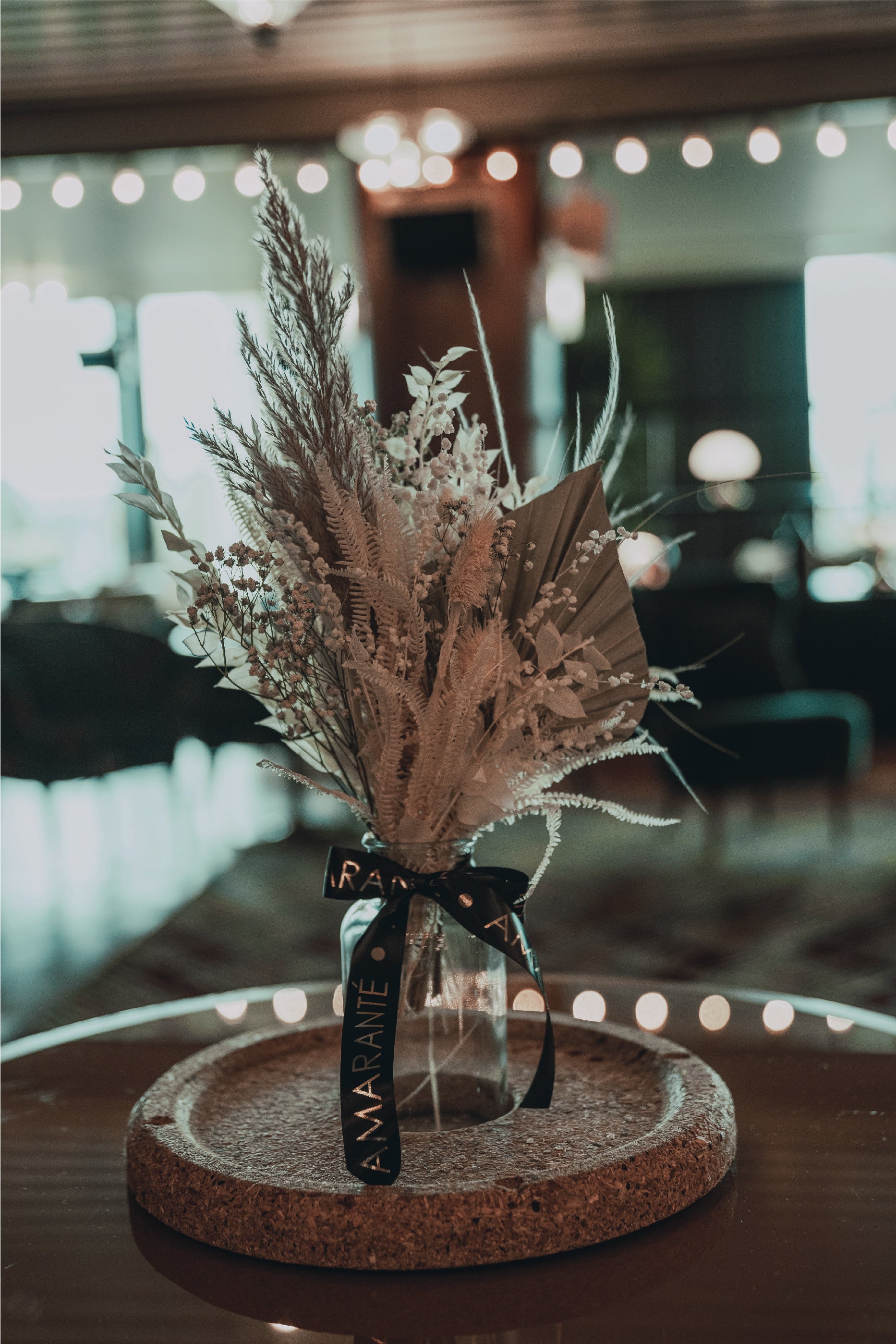 An Amaranté designed bespoke floral arrangement is showcased in this image, with an ethereal collection of white and pale green foliage accented by darker leaves, elegantly tied with a black ribbon around the glass vase, set upon a cork base with a softly lit, upscale lounge atmosphere in the background.