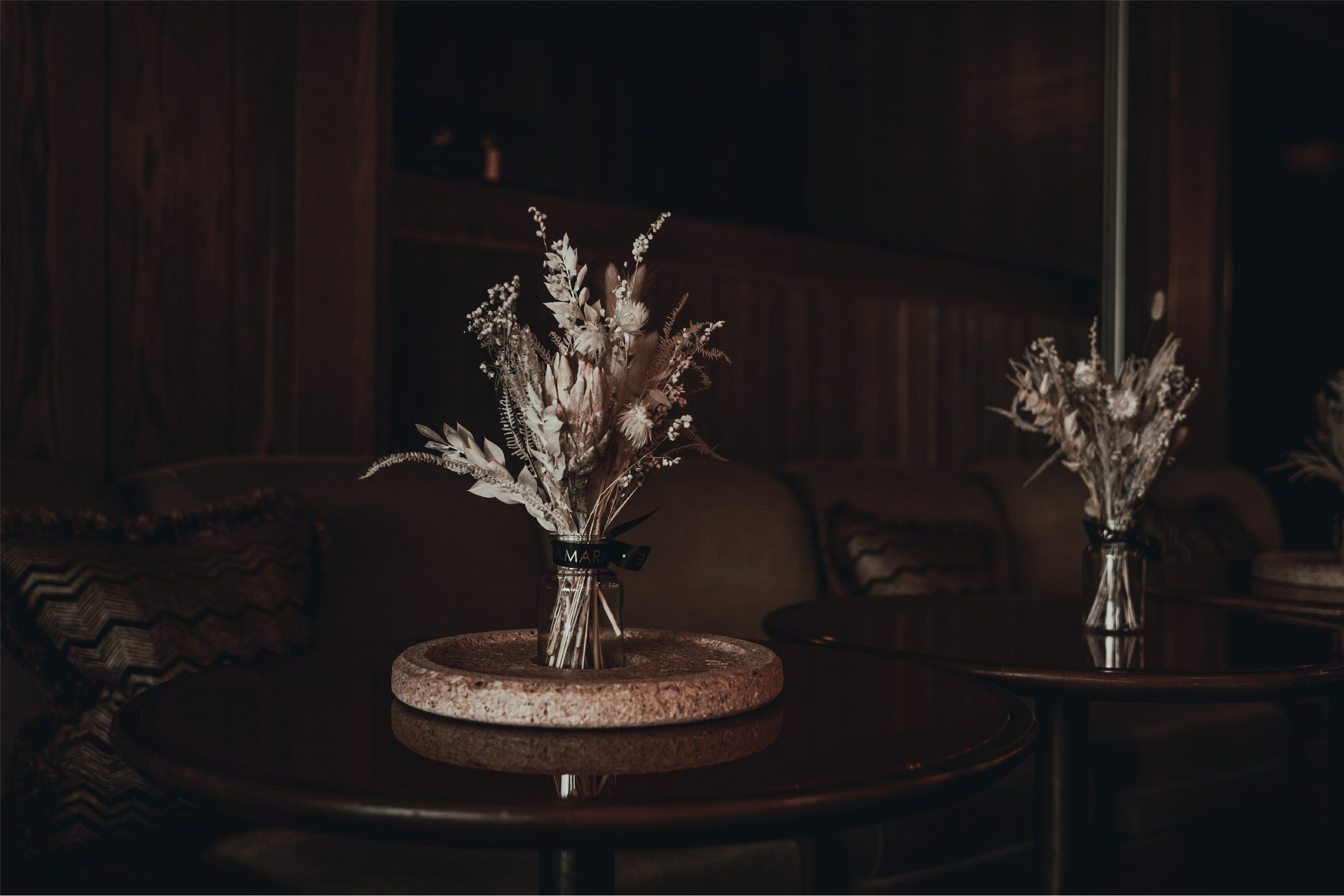 A pair of Amaranté's bespoke floral arrangements for this hotel space gracing a sophisticated venue with delicate dried botanicals in neutral tones that emanate a sense of timeless elegance, set upon circular cork bases on polished wood tables in the dimly lit, intimate ambience of a private intimate space.