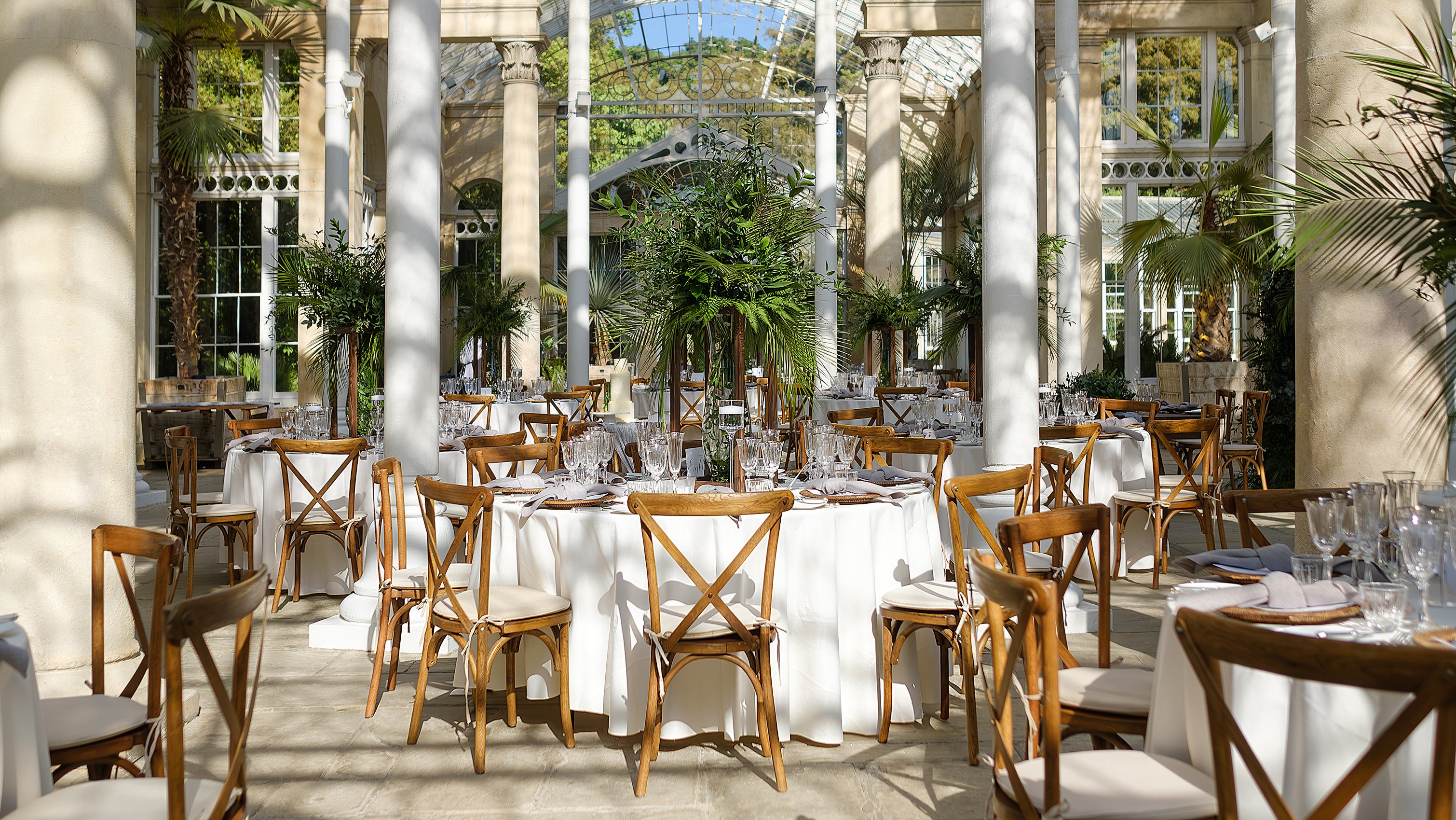 Wedding reception setup at Syon Park conservatory with wooden cross-back chairs, white linens, and tropical plants among classical columns - Floral Design  by Amaranté London