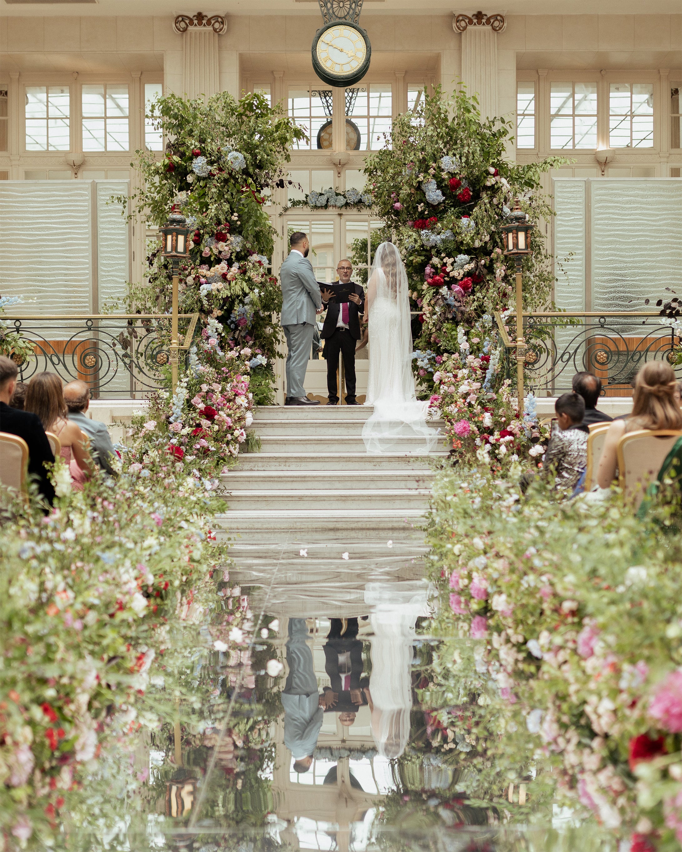 Garden-inspired wedding ceremony at Waldorf Hilton Palm Court with lush floral arch featuring red roses, pink blooms, and trailing greenery reflected in mirrored aisle - Amaranté London.