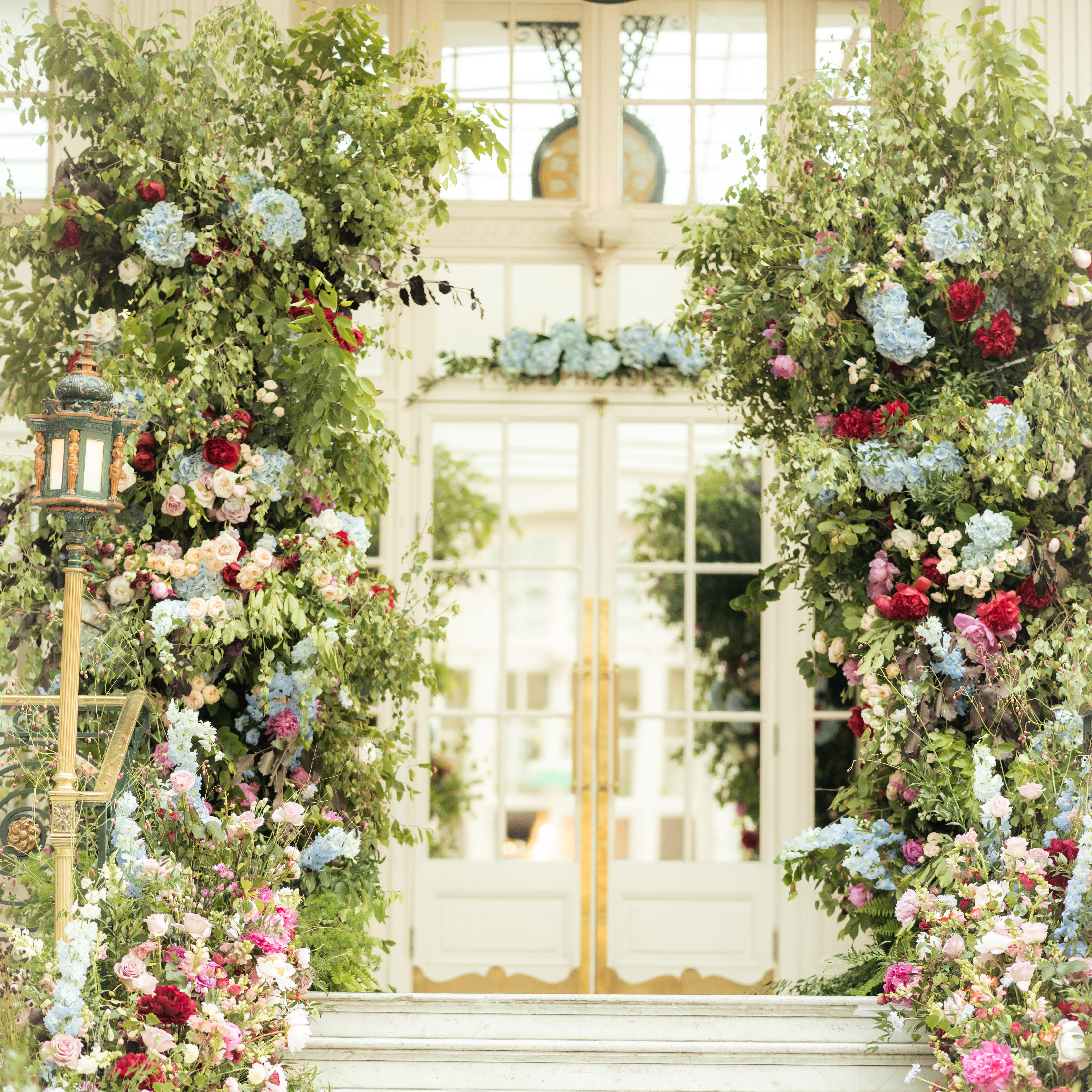 Dramatic floral ceremony arch at the Waldorf Hilton, featuring abundant trailing greenery, red peonies, blue hydrangeas, pink roses, and white blooms, creating an enchanting garden entrance - Amaranté London.