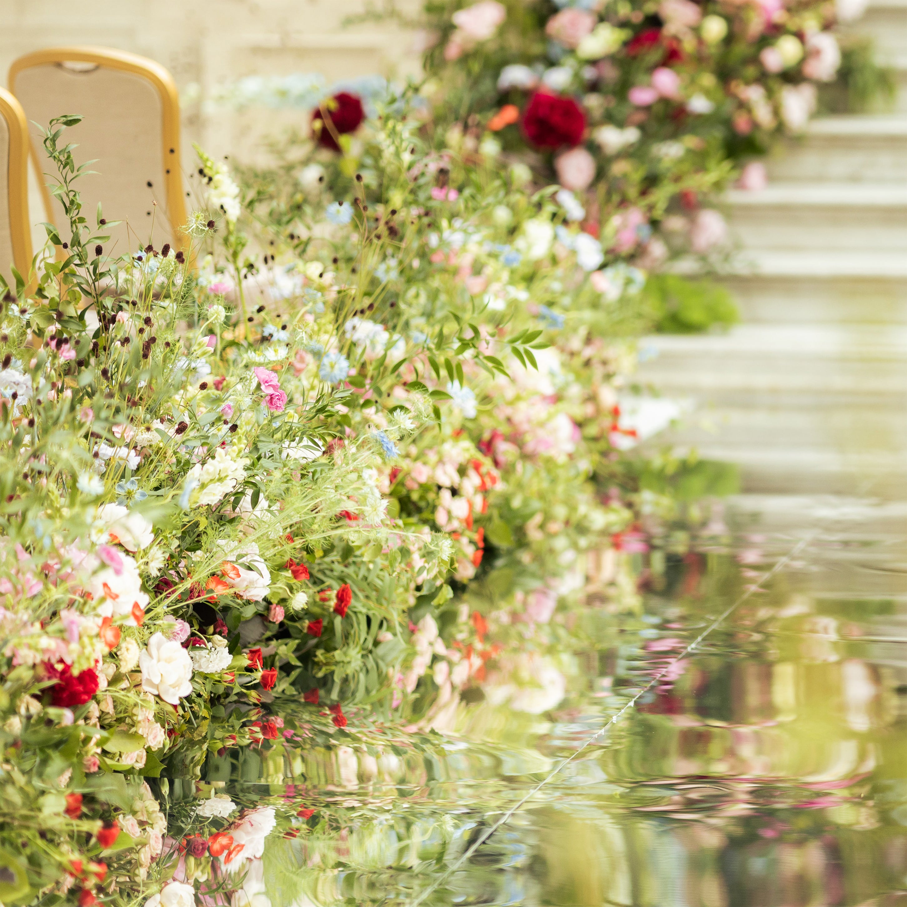 Whimsical meadow-style floral aisle arrangements at Waldorf Hilton with wildflowers, red dahlias, pink roses, blue delphiniums, and ferns reflected in a mirrored surface - Amaranté London.