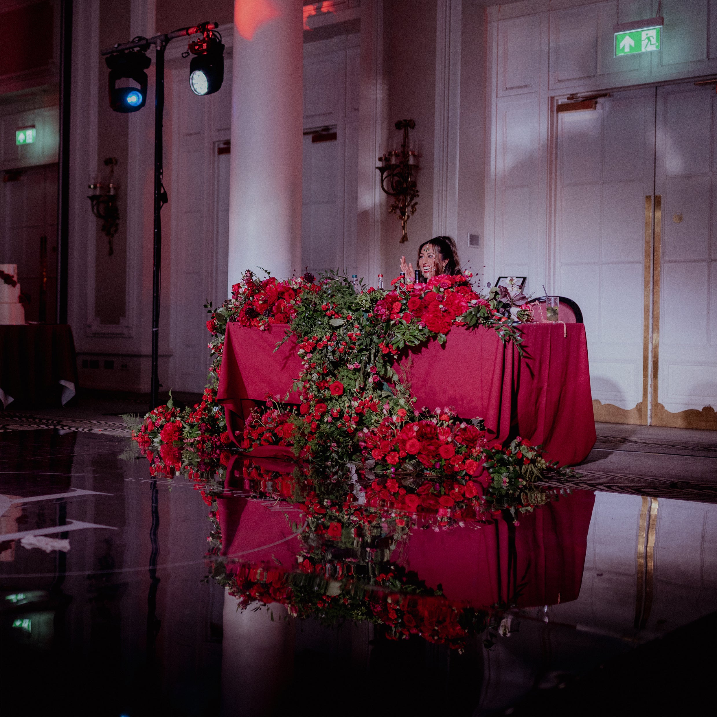 Dramatic cascading red floral garland at Waldorf Hilton with red roses and greenery adorning the DJ booth, reflected in the polished floor with atmospheric pink uplighting - Amaranté London.