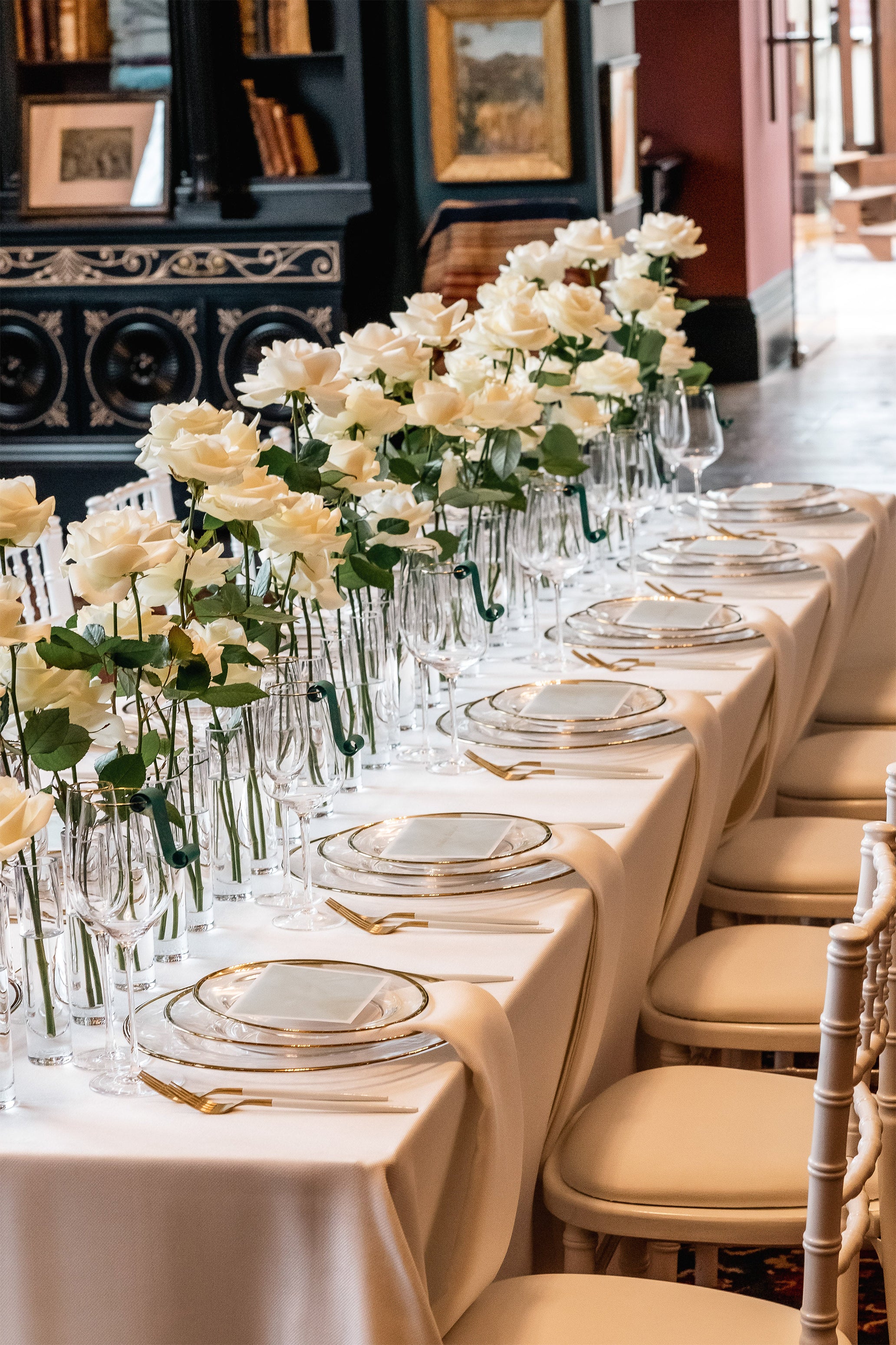 Elegant long dining table with repeated white rose arrangements in crystal vases, creating floral rhythm with fine china and crystal glassware - Floral Design by Amaranté London