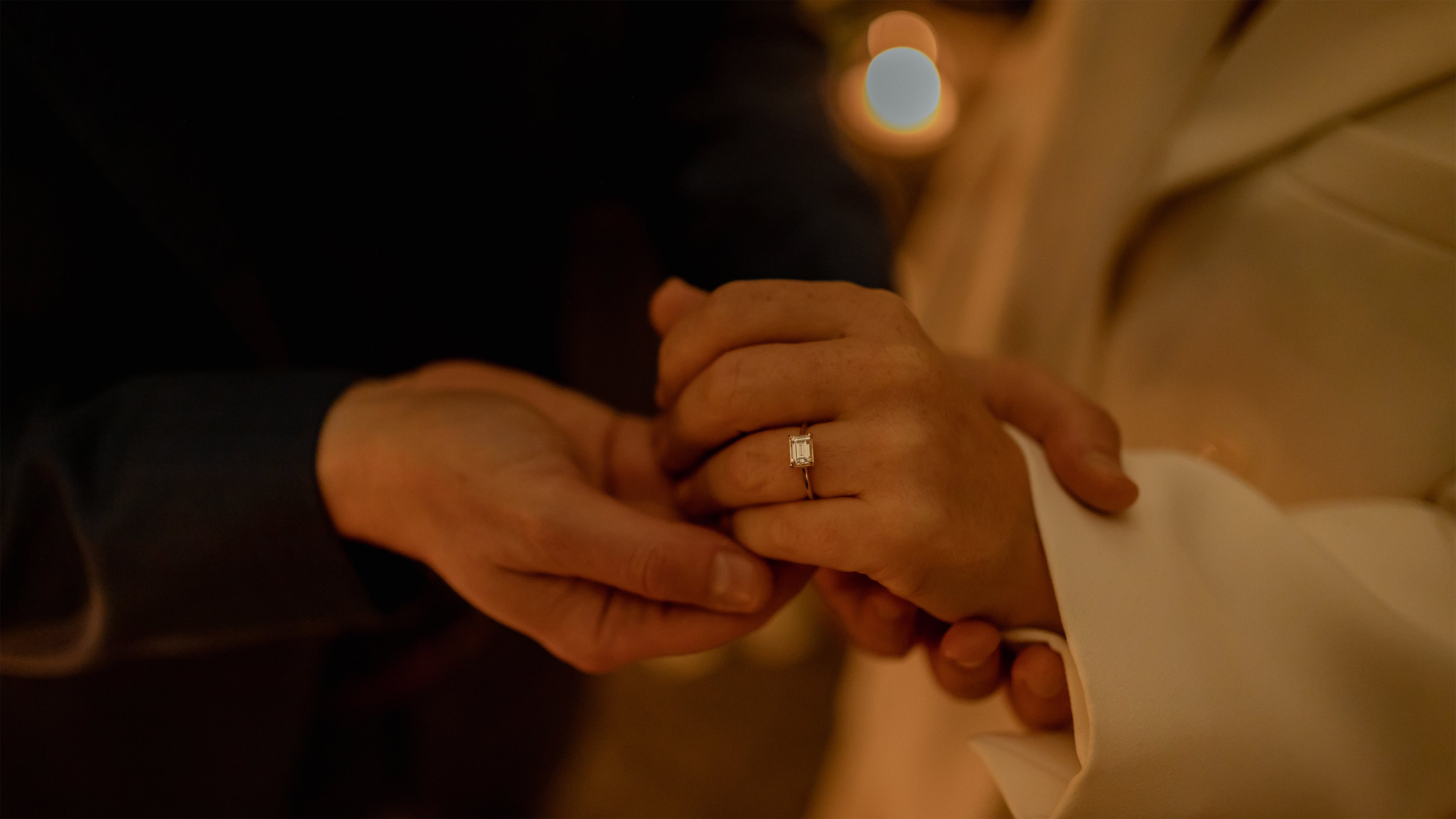 ALT: "Couple holding hands showing emerald-cut diamond engagement ring in warm candlelight after romantic proposal - Amaranté London