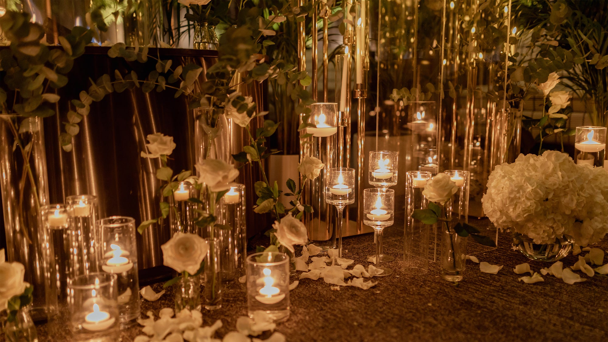 Romantic proposal setup with floating candles in glass cylinders, white roses, eucalyptus garland, and scattered rose petals creating intimate ambiance - Amaranté London