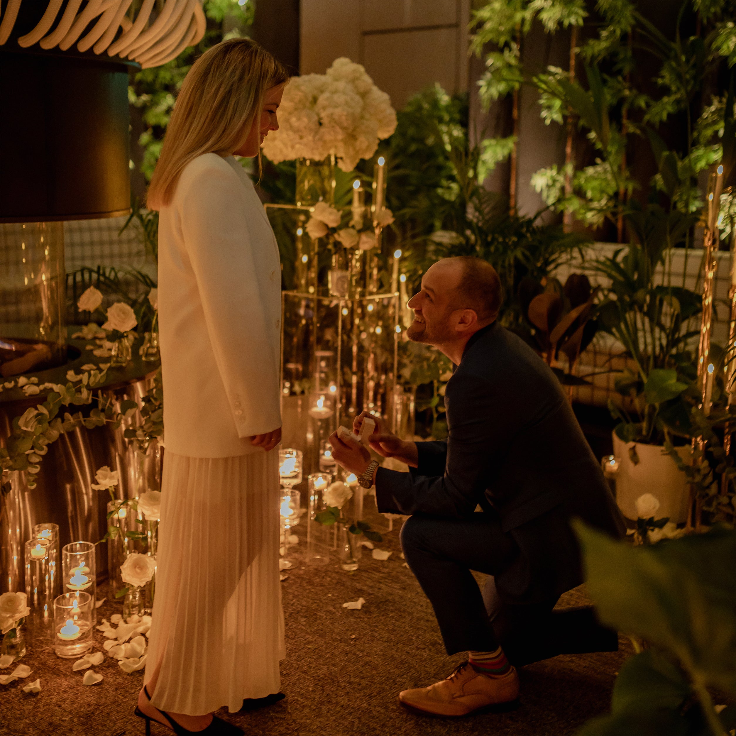 Marriage proposal moment with man on bended knee presenting ring, surrounded by white hydrangea centerpieces, floating candles, and lush greenery installation - Amaranté London
