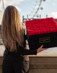 A woman elegantly dressed with long blond hair, seen from behind, holding a large square black suede box filled with lush red infinity roses, with the London Eye in the background - Amaranté London.